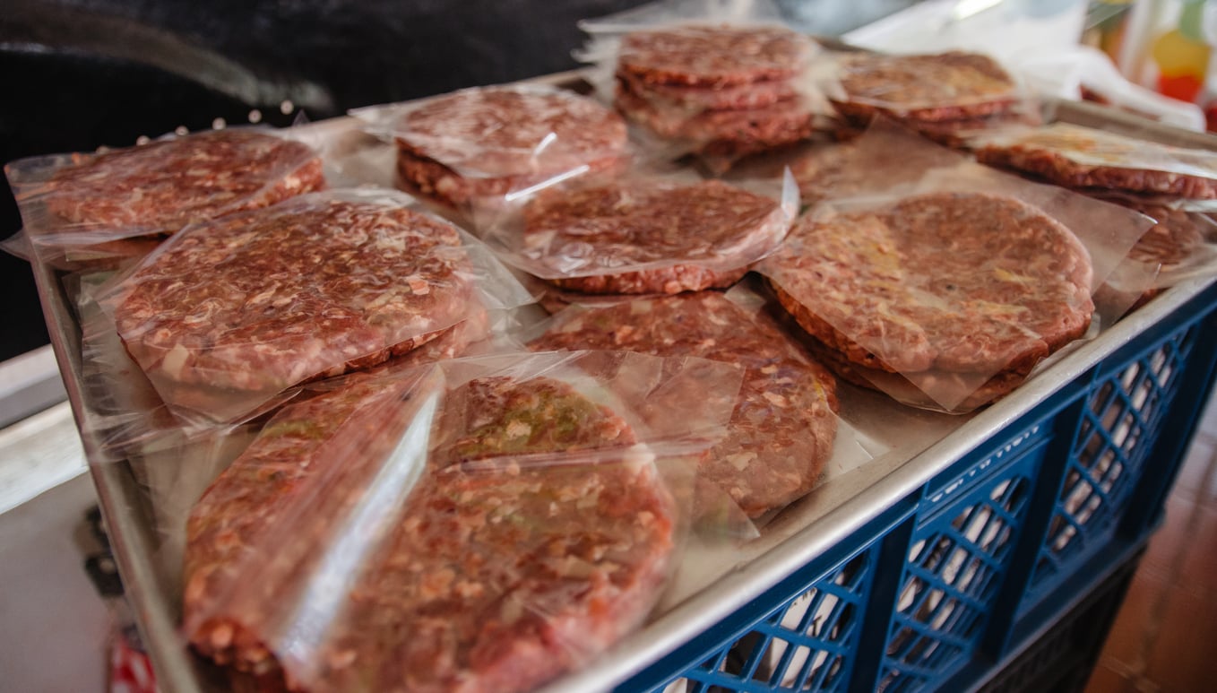 Raw hamburger patties individually wrapped in plastic sit on a metal tray atop a blue crate, ready for cooking or freezing.