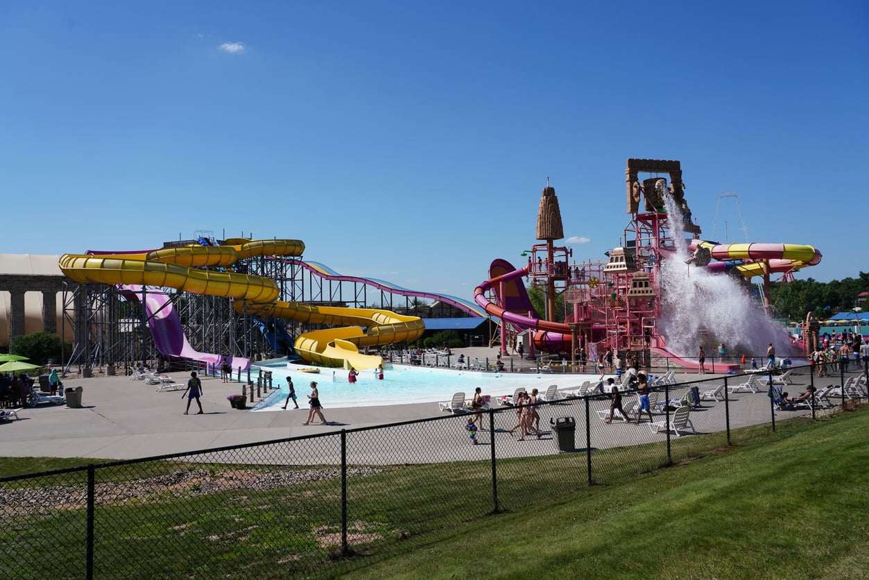 A water park with colorful slides, a large splash structure, people swimming and lounging, and a big bucket of water dumping onto the play area under a clear blue sky—one of the most exciting affordable travel destinations for family fun.