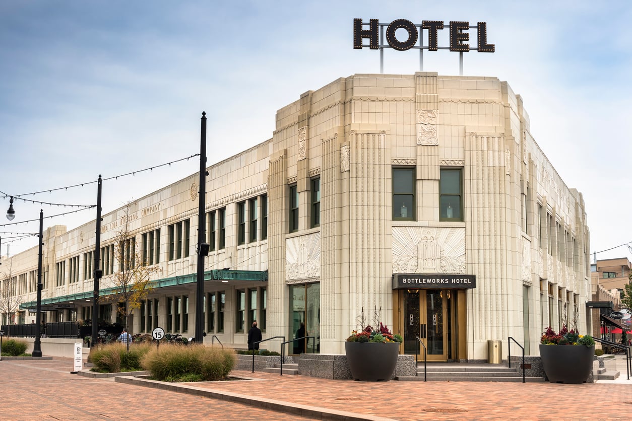 Art Deco style white building with large "HOTEL" sign on roof, labeled "Bottleworks Hotel" above entrance. Wide sidewalks, potted plants, and string lights line the street. People walk nearby under a cloudy sky, perhaps eager to check in and avoid common hotel booking mistakes as they arrive at this stylish destination.