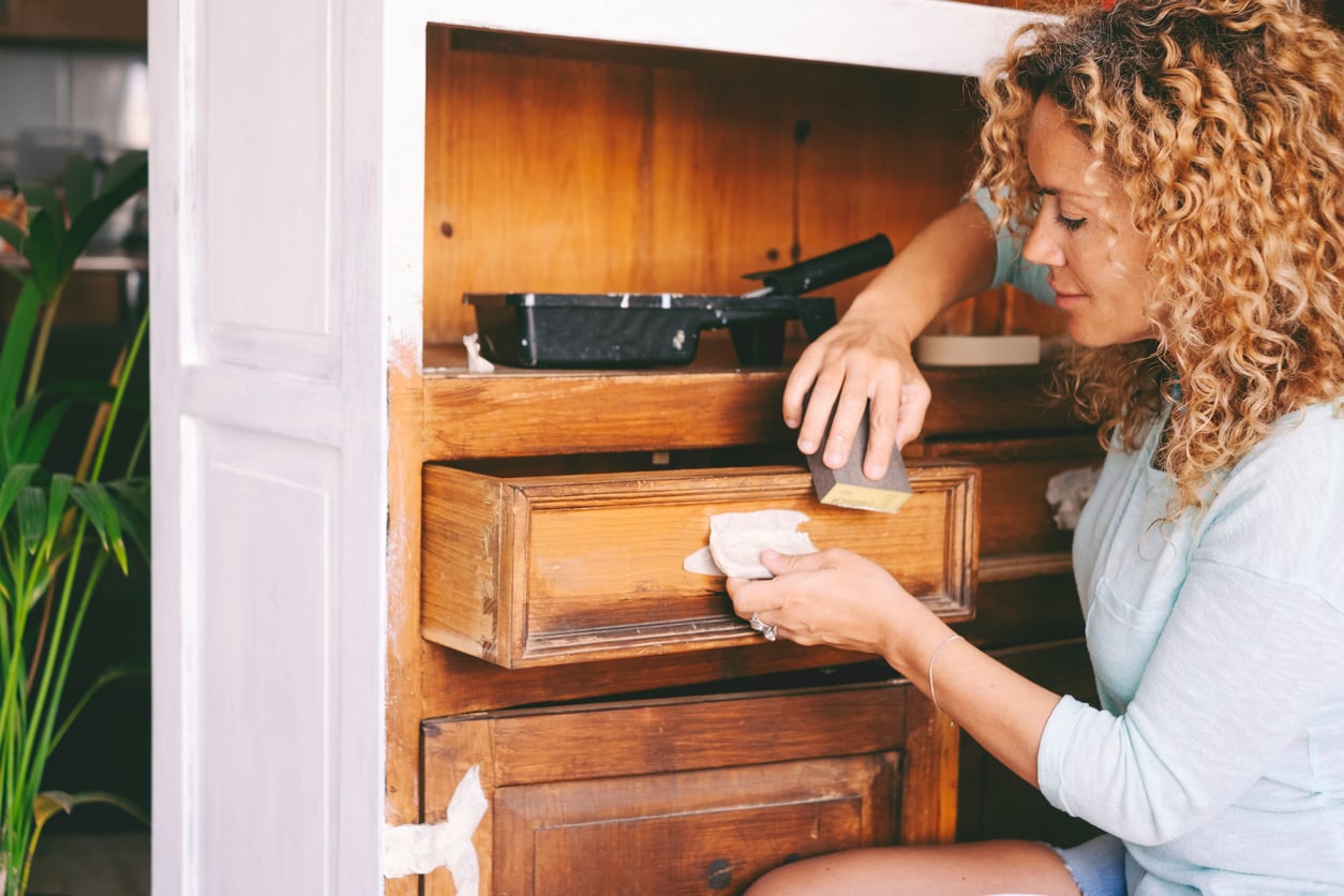 A woman with curly hair sands a wooden drawer while working on refinishing a wooden cabinet. A paint tray is on top of the cabinet, and a green plant is visible in the background.