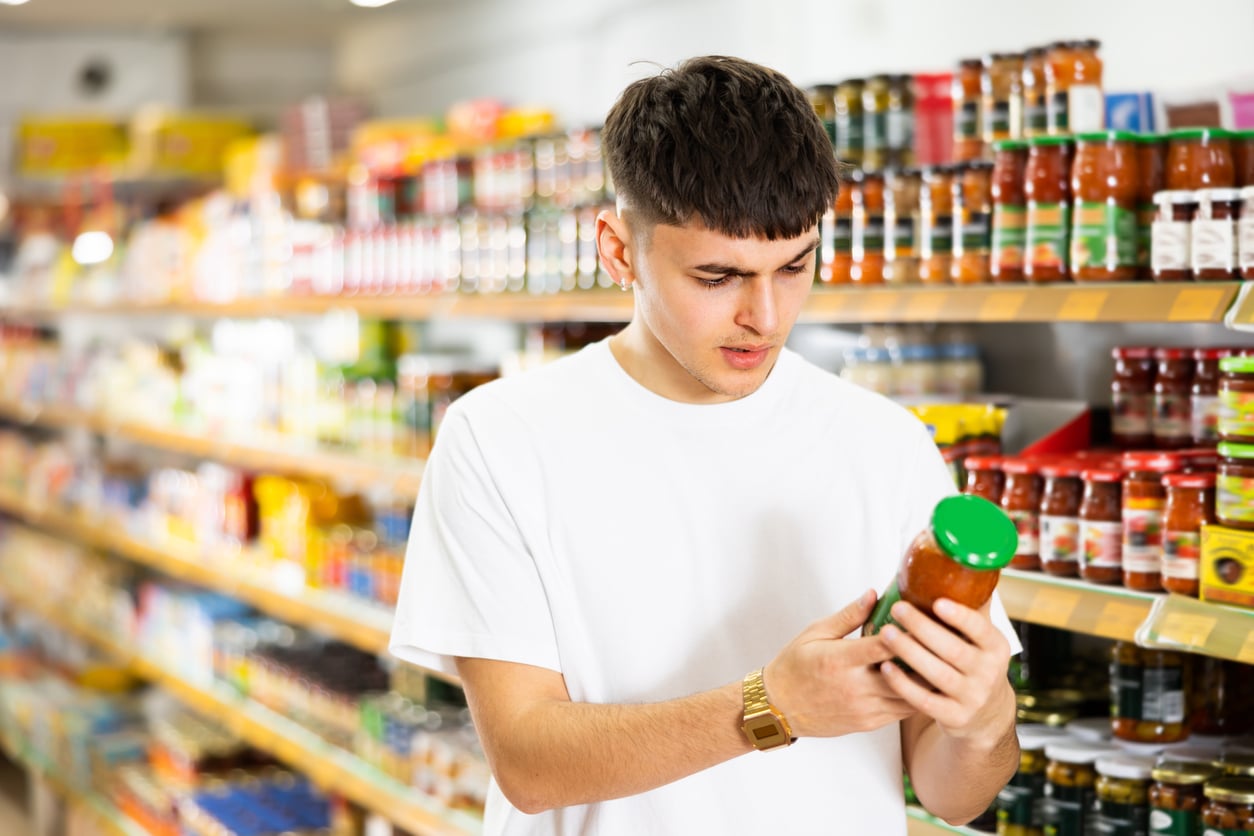 A young man in a white t-shirt stands in a grocery store aisle, closely examining a jar of sauce or condiment with a green lid, demonstrating his money saving habits as he carefully compares options amidst shelves filled with various products.