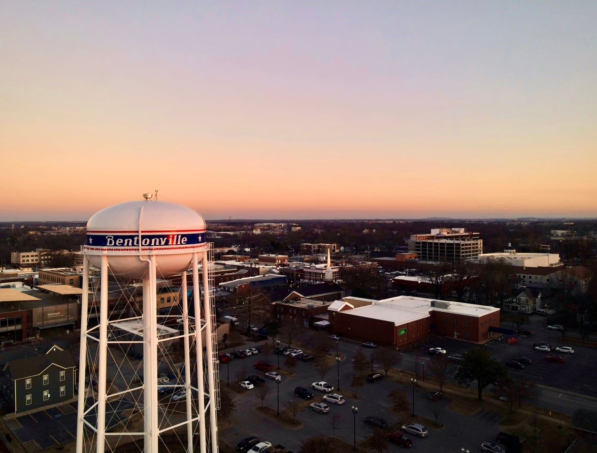 A water tower labeled "Bentonville" stands over a town at sunset, with buildings, parked cars, and a colorful sky in the background—making it one of the charming travel destinations to explore.