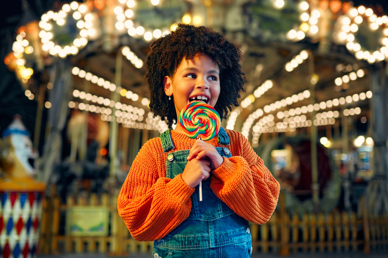 A smiling child with curly hair holds a large colorful lollipop while standing in front of a brightly lit carousel at an amusement park.