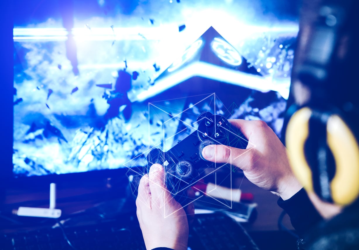 Person wearing headphones plays a video game using a controller in front of a computer monitor displaying a futuristic, blue sci-fi scene with glowing geometric shapes.