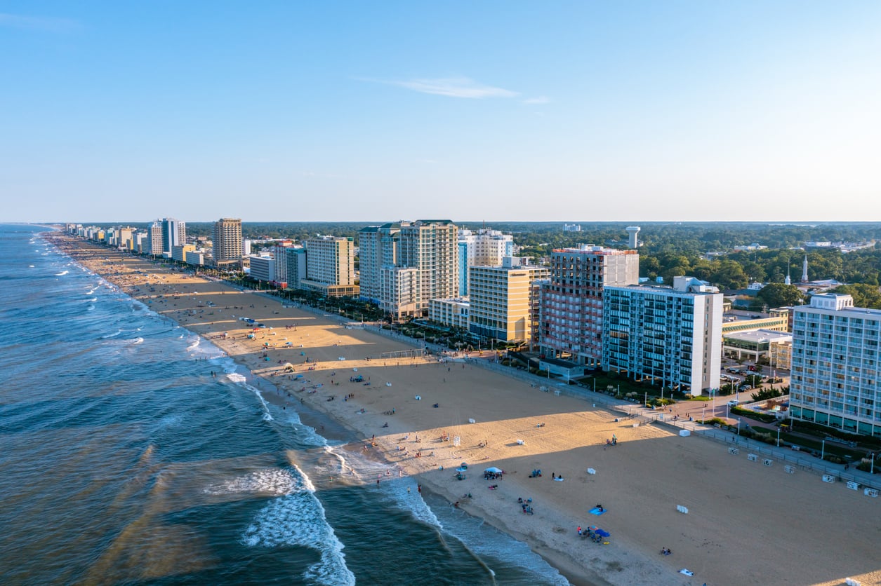 Aerial view of a sandy beach lined with tall hotels and buildings, with waves gently hitting the shore and people scattered along the sand on a clear, sunny day.