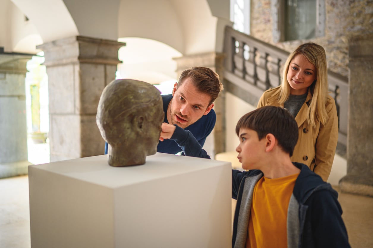 A man, a woman, and a boy observe a sculpted head on a pedestal in a museum. The man and boy examine it closely while the woman smiles in the background, perhaps reflecting on how museum visits are one of those memberships worth paying for. Stone columns and stairs are visible behind them.