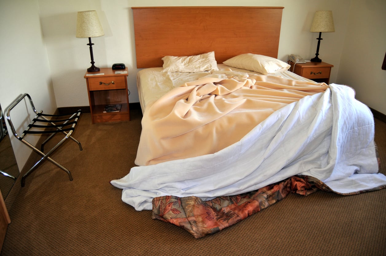 A bed with rumpled sheets and blankets partially on the floor, flanked by two nightstands with lamps, in a simply furnished room—an all-too-familiar sight for travelers who've experienced hotel booking mistakes.