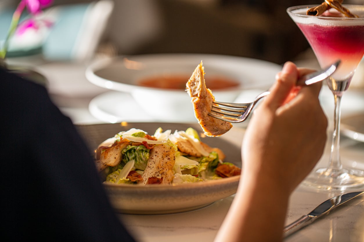 A person holds a fork with a piece of chicken over a Caesar salad, perhaps savoring the flavors while calculating vacation costs. A pink cocktail and a bowl of soup are visible on the table in a well-lit restaurant setting.