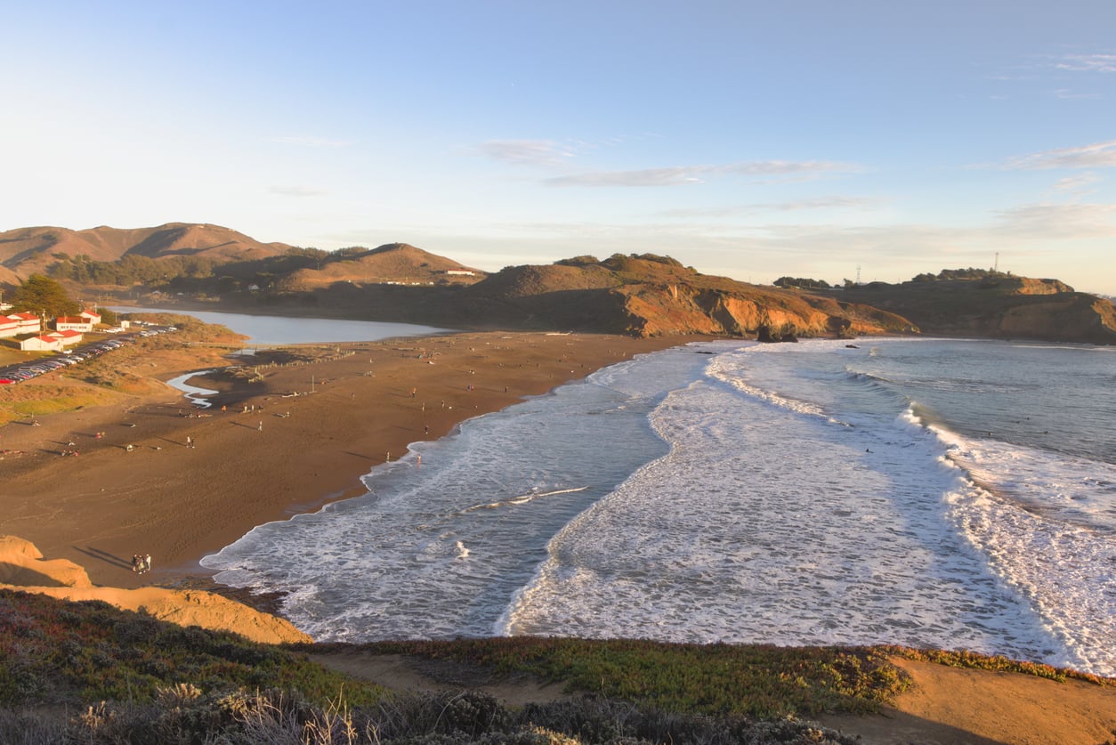 View of a sandy beach with gentle waves, rugged hills in the background, and scattered people walking along the shore under a clear sky in warm, golden light—a classic scene among travel destinations on the West Coast.