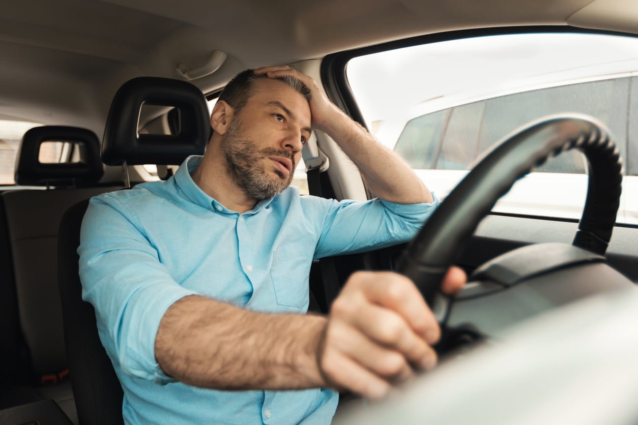 A man in a light blue shirt sits in a car, looking tired and stressed with his hand on his forehead and leaning on the steering wheel, perhaps weighed down by worries about money saving habits.