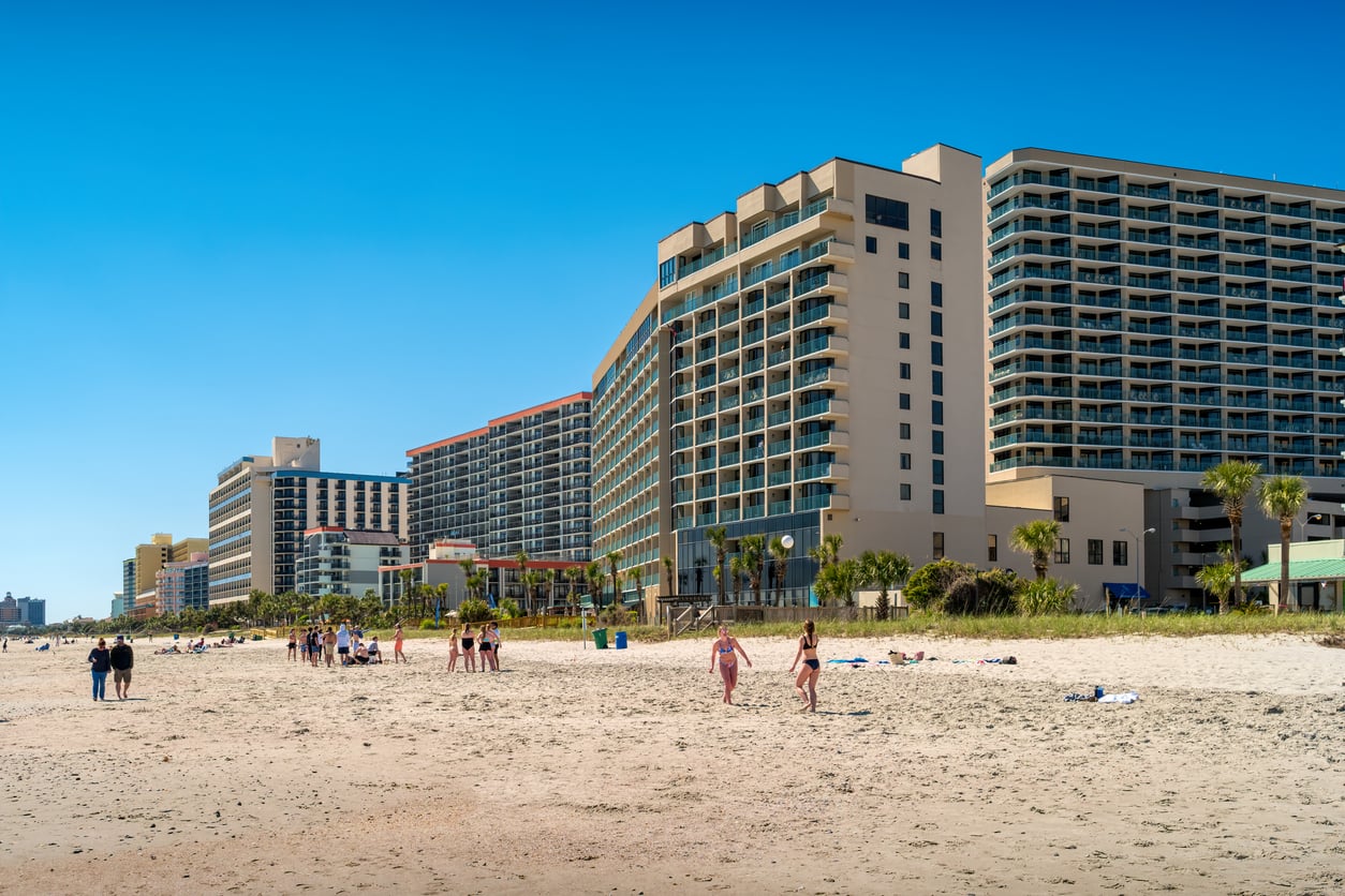 People walk and relax on a sandy beach in front of several large beachfront hotels under a clear blue sky. Palm trees line the area between the hotels and the beach, making it a perfect spot to enjoy your beach vacation savings.
