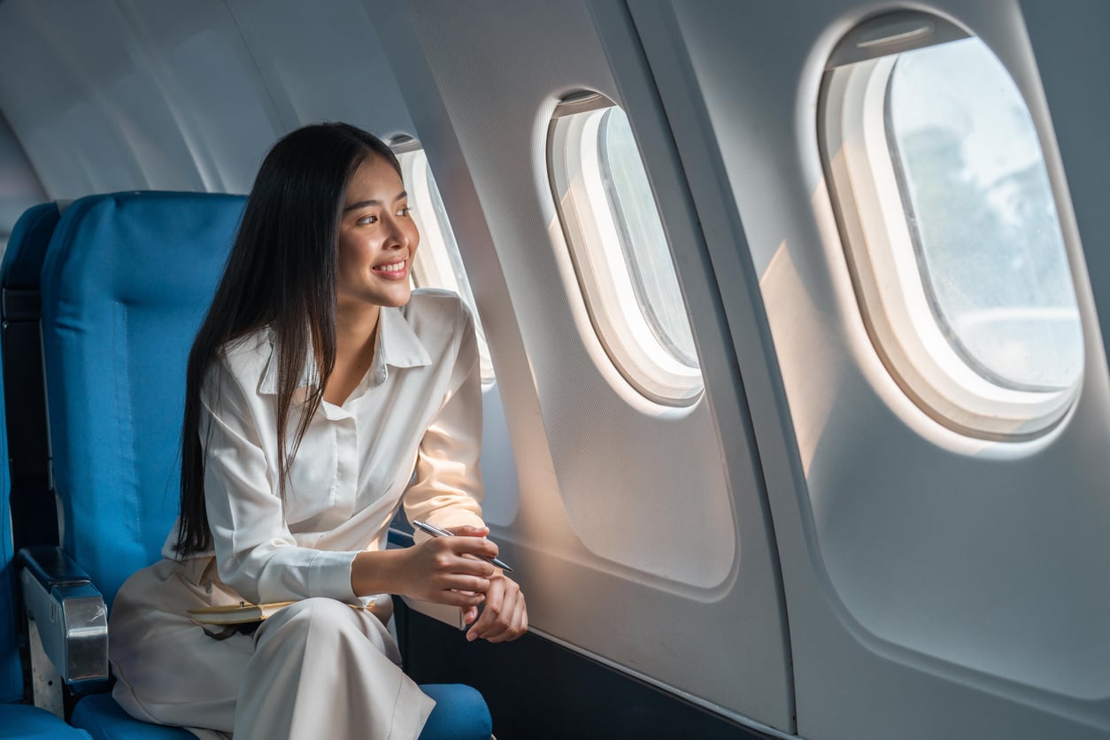 A woman sits in an airplane seat next to the window, smiling and looking outside. She is wearing a white outfit and holding a notebook or folder in her hands. Sunlight shines through the windows onto her face.
