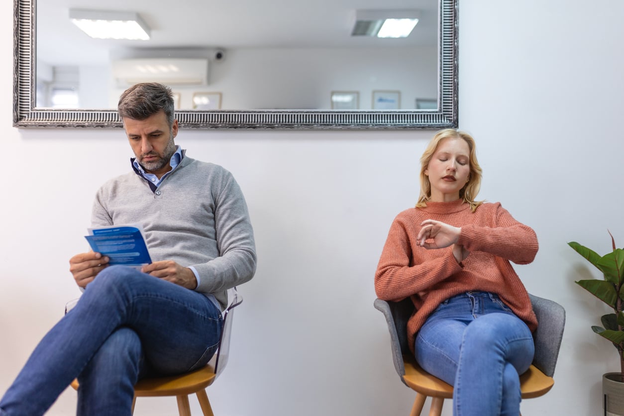 A man reads a pamphlet and a woman checks her watch as they sit in chairs in a waiting room with a large mirror behind them, capturing a glimpse of life before smartphones.
