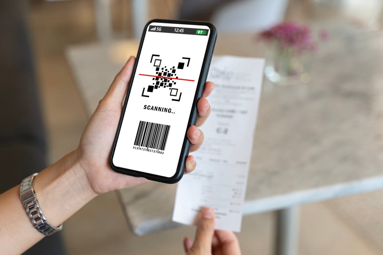A person holds a smartphone scanning a QR code on a receipt in a cafe. The phone screen shows a scanning progress bar and barcode below the QR code.