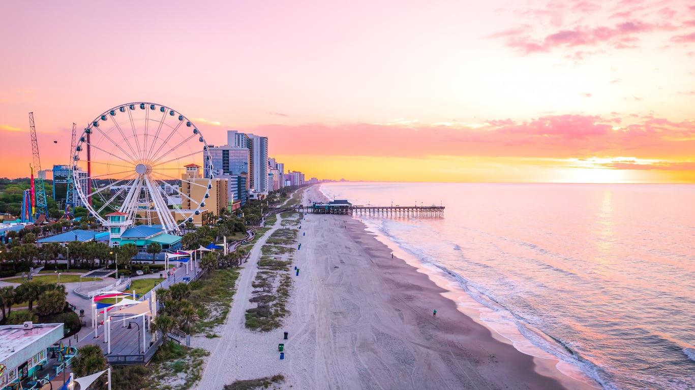 Aerial view of an East Coast beach at sunrise with a Ferris wheel, pier, and buildings along the shoreline. The sky is pink and orange, reflecting on the calm ocean waves.