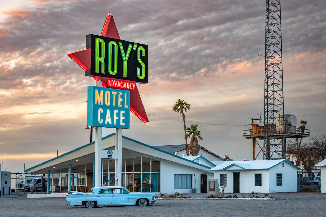 A vintage blue car parked in front of Roy's Motel and Café sign, with a retro building, palm trees, and a cloudy sunset sky capturing the nostalgic charm of classic American towns in the background.