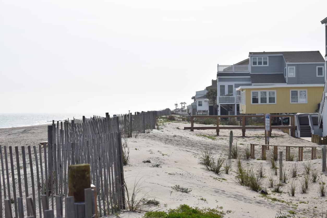 Beachfront houses stand beside a sandy shore lined with a wooden fence and sparse vegetation, evoking the quiet charm of classic beach towns. The sky is overcast, and the ocean is visible on the left side of the image.