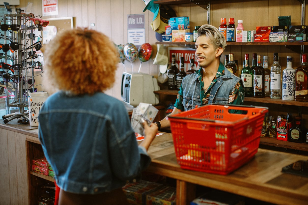 A person with curly hair hands cash to a smiling cashier at a small store counter, with a red shopping basket and shelves of various products and liquor bottles behind them.