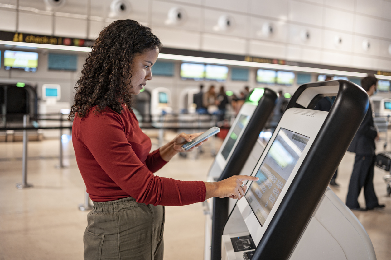 A woman uses a self-service kiosk at an airport check-in area, holding her phone in one hand and touching the screen with the other. Digital screens and check-in counters are visible in the background.