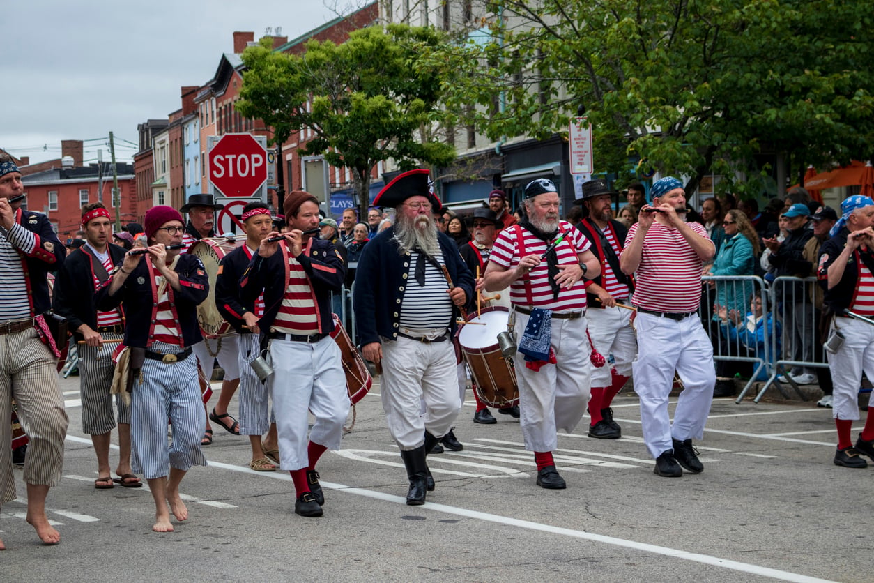 A parade features people in red, white, and blue outfits, playing drums and flutes while marching down a street lined with spectators, creating a festive atmosphere reminiscent of Halloween celebrations.