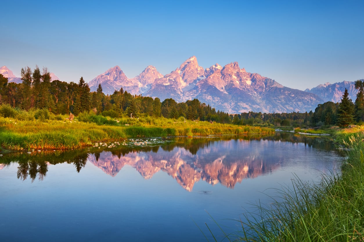 A calm river reflects tall, rugged mountains and green trees under a clear blue sky, with soft sunlight illuminating the landscape.