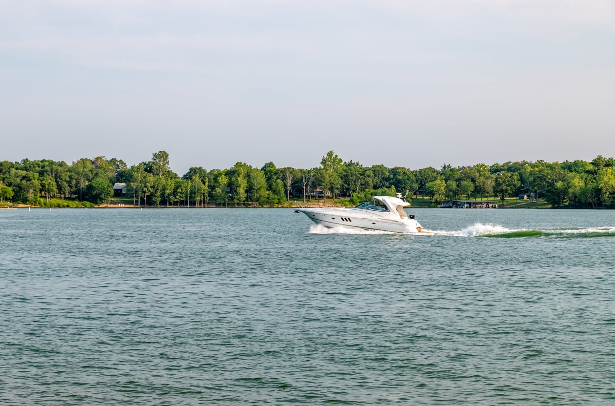 A white motorboat speeds across a wide lake with green trees lining the opposite shore under a partly cloudy sky.