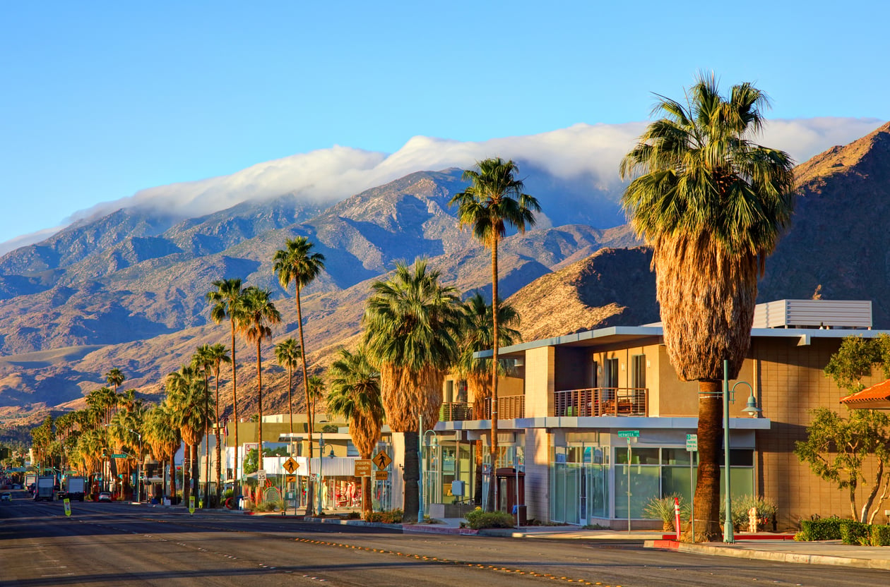 A sunny street lined with palm trees and modern buildings, with rugged mountains and a layer of clouds in the background under a clear blue sky—one of the most captivating travel destinations for those seeking both relaxation and adventure.