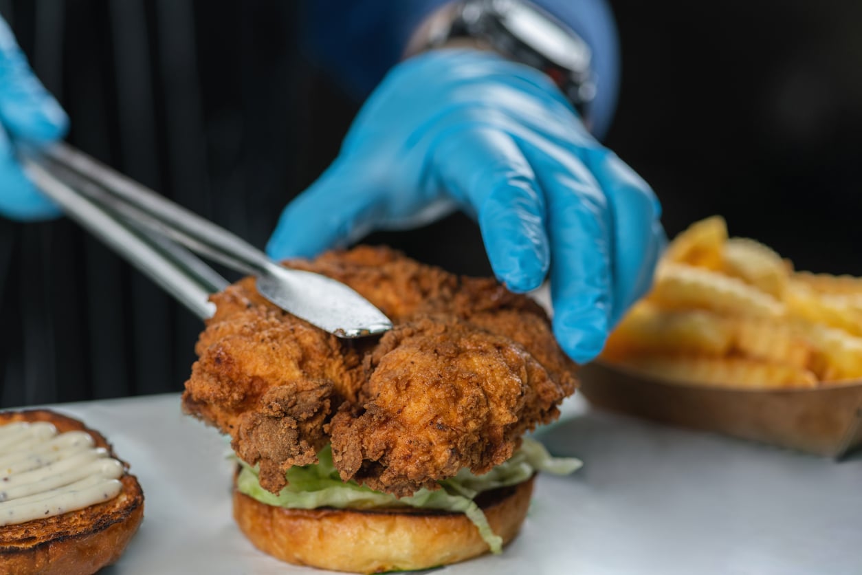 A person wearing blue gloves uses tongs to place fried chicken on a sandwich bun with lettuce and mayonnaise. Crinkle-cut fries are visible in the background.