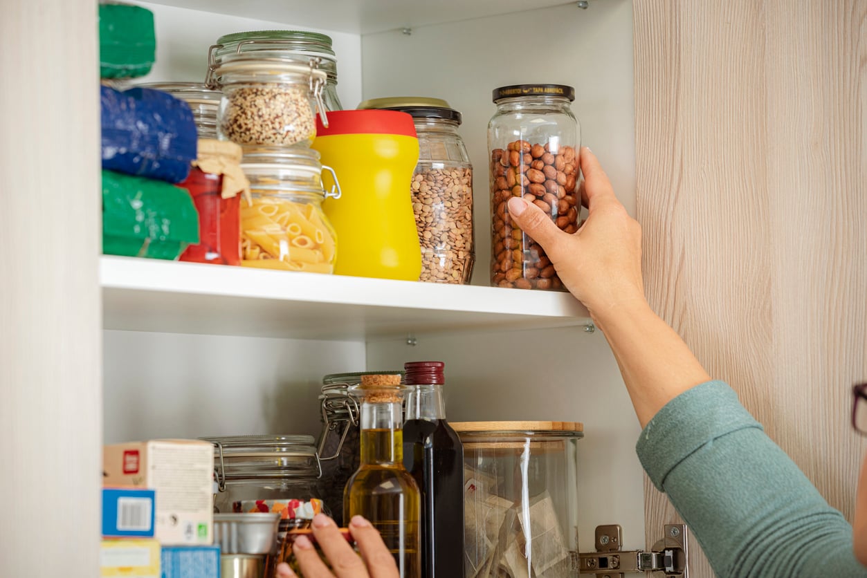 A person reaches for a jar of beans on a pantry shelf filled with jars of grains, pasta, and bottles of oil—each item neatly arranged as a reflection of thoughtful grocery shopping habits. Their other hand is near items on the lower shelf. The pantry shelves are white inside a wooden cabinet.