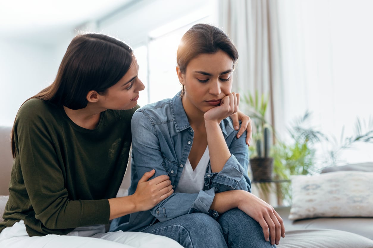 A woman comforts a sad friend by sitting close and placing an arm around her shoulder on a couch in a bright living room.