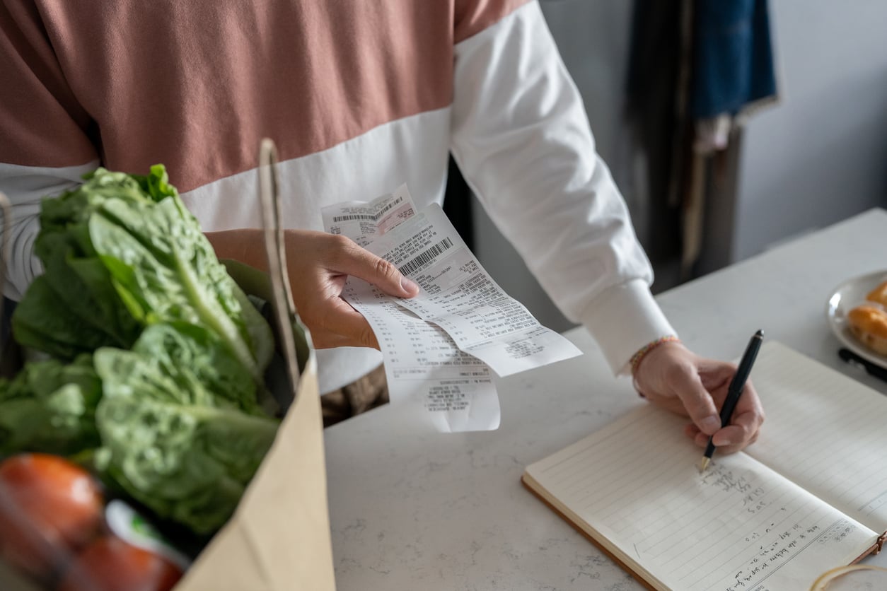 A person holds several receipts next to a grocery bag filled with fresh produce, while writing or calculating expenses in an open notebook on a white countertop—a reflection of how families in America are having fewer children.