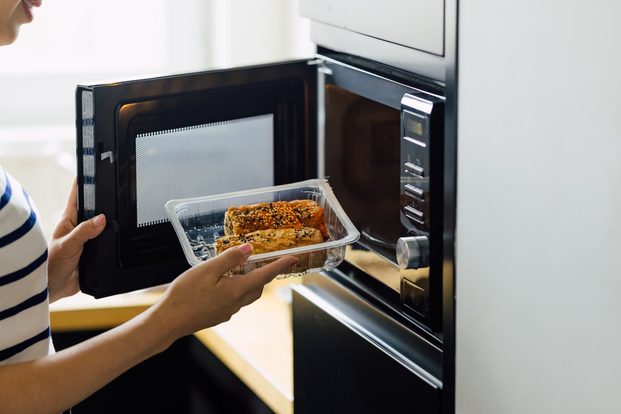 A person in a striped shirt puts a plastic container with food into a microwave oven, preparing to heat the meal.