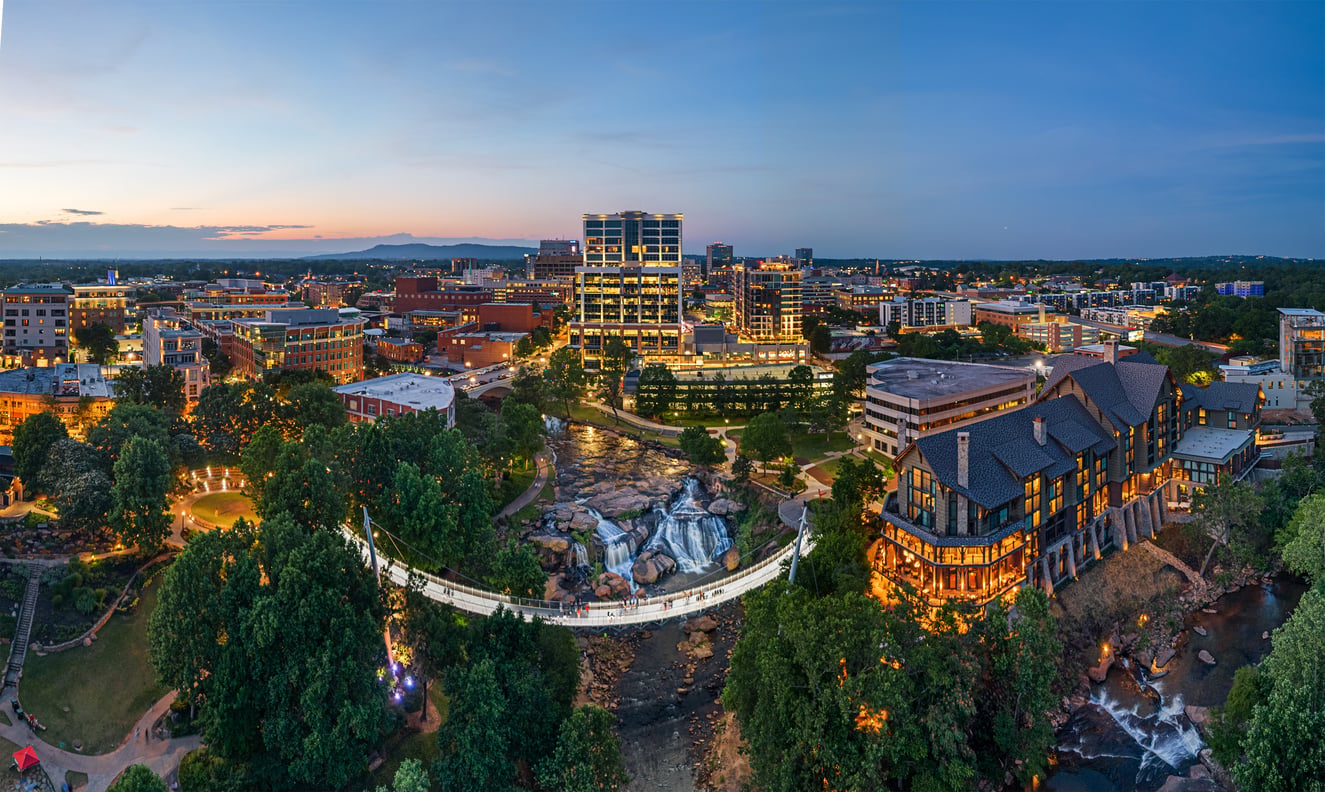 Aerial view of a cityscape at dusk with illuminated buildings, a pedestrian suspension bridge over a river, and a park surrounded by trees and greenery—one of the 20 travel destinations that offer an unforgettable urban adventure paired with natural beauty.