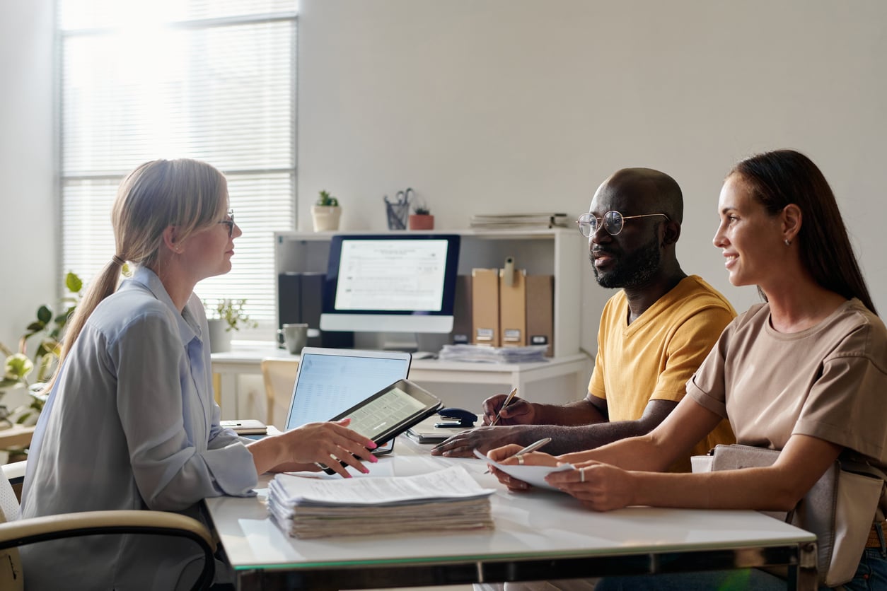 Three people sit at a desk in an office, engaged in a discussion. One woman faces a man and another woman, both holding papers; computers and documents are visible in the background.