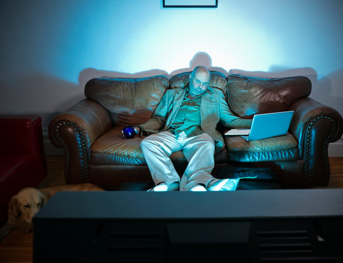 A man in a suit naps on a brown leather couch with a laptop on his lap and a TV remote in his hand, illuminated by the blue glow from a television. A dog lies on the floor nearby.