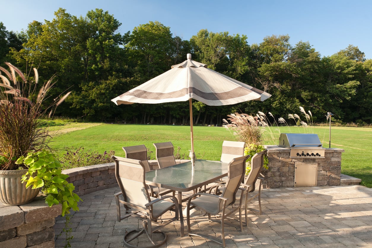 Patio with a glass table, six chairs, and a large umbrella. A stainless steel grill sits nearby alongside premium outdoor home products, with grass, trees, and a stone patio, all under a clear blue sky.