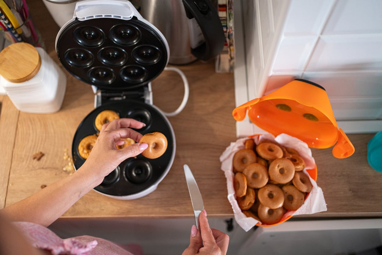 A person uses a knife to remove mini donuts from a donut maker—one of their favorite kitchen gadgets—on a kitchen counter, with a bowl of finished mini donuts nearby.