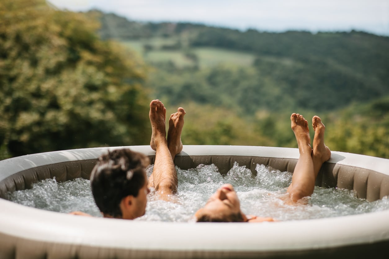 Two people relax in an outdoor hot tub with bubbling water, surrounded by lush greenery and distant hills under a cloudy sky. Their legs are stretched out above the water, and their faces are partially visible, enjoying one of the finest outdoor home products for unwinding in nature.