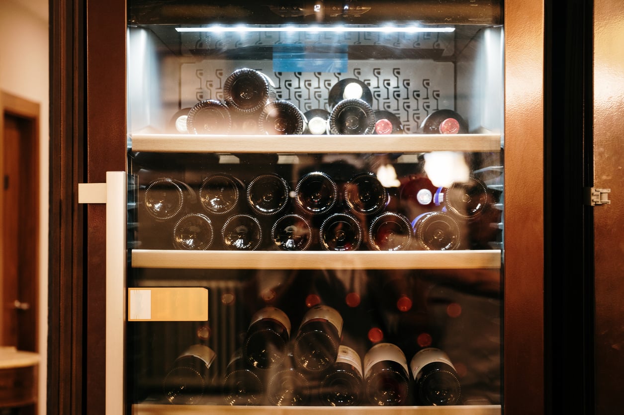 A wine fridge filled with several rows of wine bottles lying horizontally on shelves, viewed through the glass door with reflections visible. The fridge is well-lit and appears to be in a modern setting.