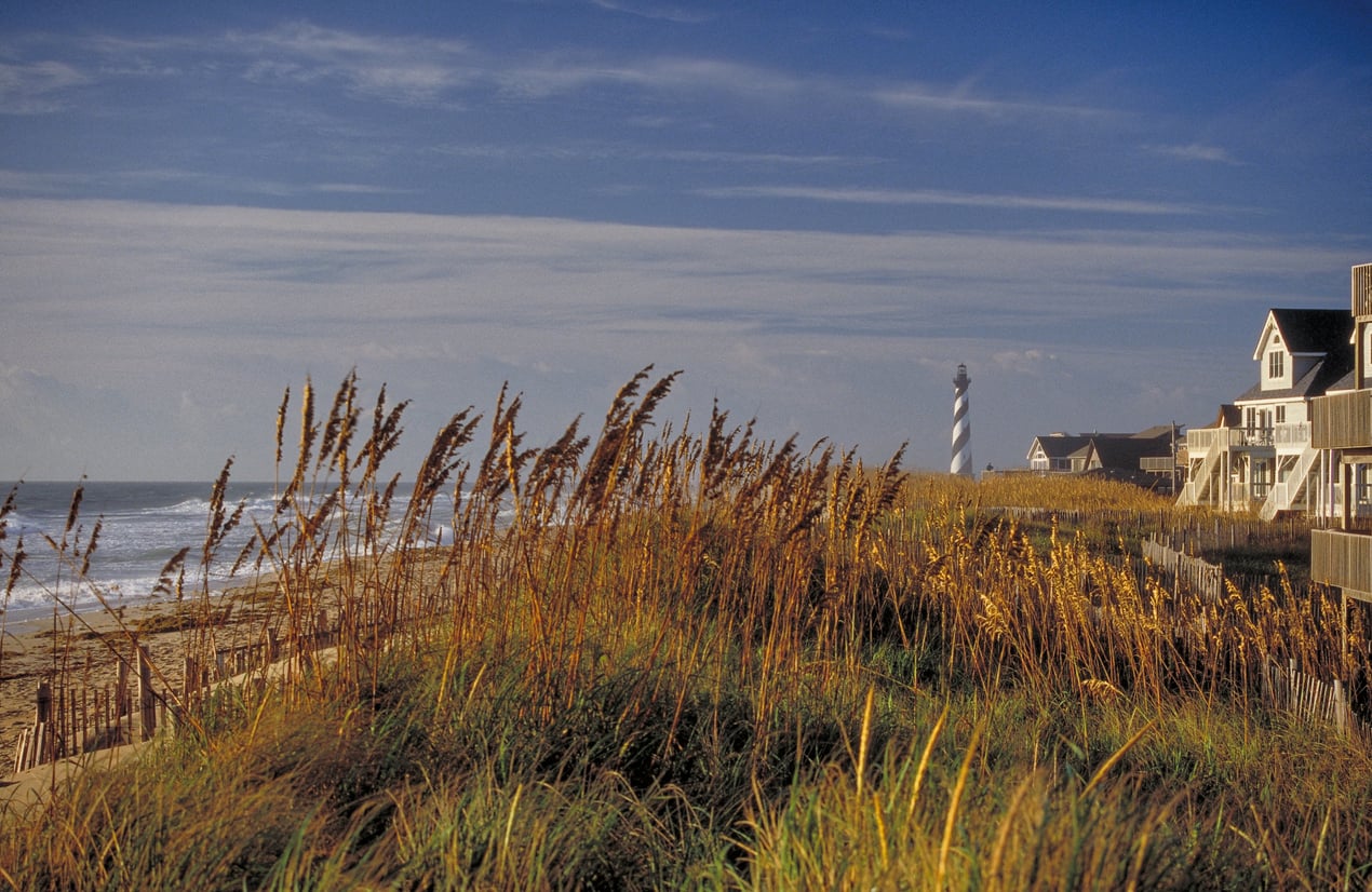 Beach houses line the right side of the image, with tall grasses in the foreground and a distant black-and-white striped lighthouse near the shoreline under a partly cloudy sky, evoking classic East Coast charm.