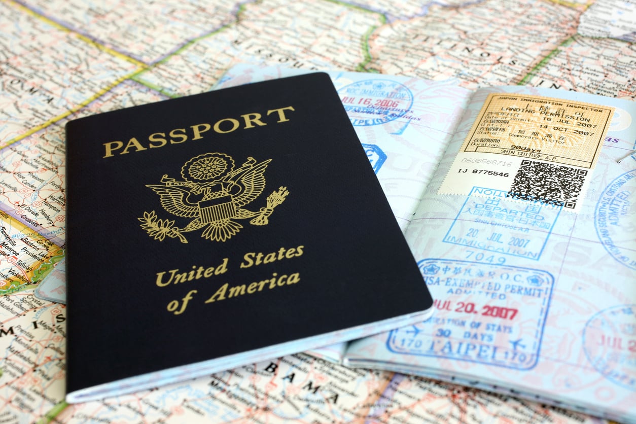 A U.S. passport rests on top of an open passport displaying various travel stamps, all placed on a detailed map—a reminder to check your documents before booking a flight.