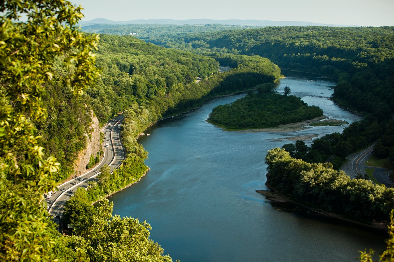 A winding river flows through a lush, green East Coast forest with a curved road running alongside it. Hills and trees stretch into the distance under a clear sky.