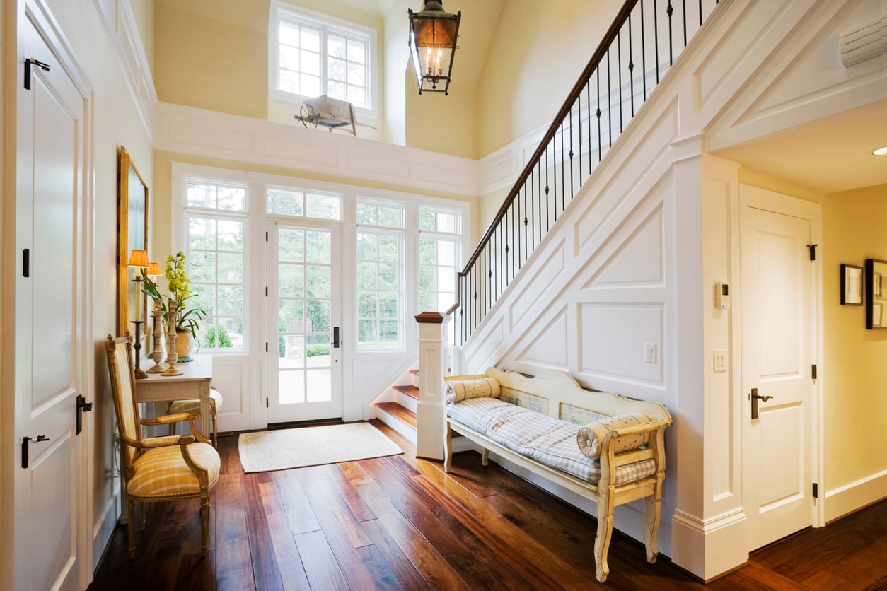Bright entryway with wooden floors, a beige upholstered bench, and a staircase with white paneling and dark railingsāunlike some home features homeowners no longer want, such as dark foyers or bulky furnishings, this space feels open and welcoming. Large windows and glass doors let in lots of natural light. A chair and console table sit against one wall.