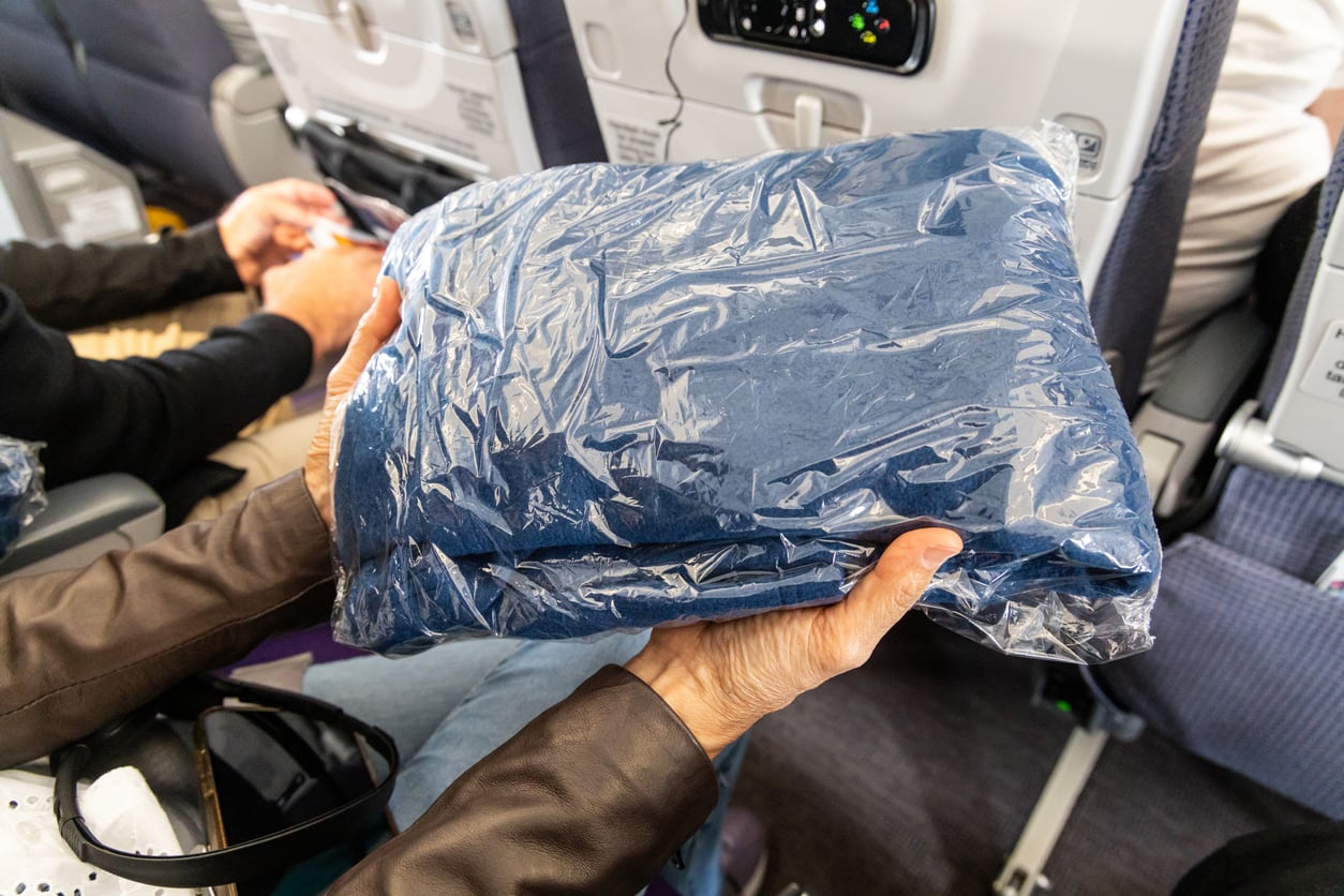 A person holds a blue airline blanket wrapped in plastic while seated on an airplane. Nearby passengers and seatback trays are visible in the background.