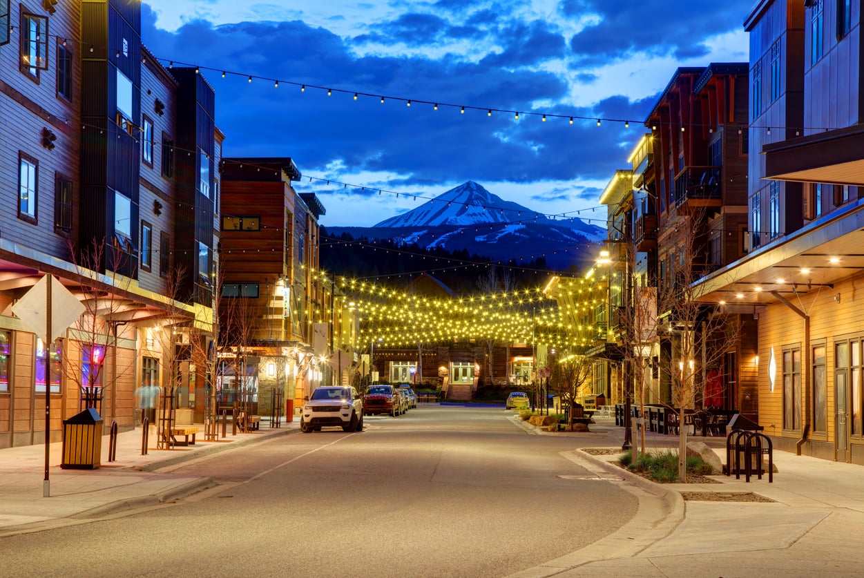 A quiet street lined with modern buildings and string lights at dusk, with a snow-capped mountain rising in the background under a blue sky, evokes the charm found in some of the world’s top 20 travel destinations.