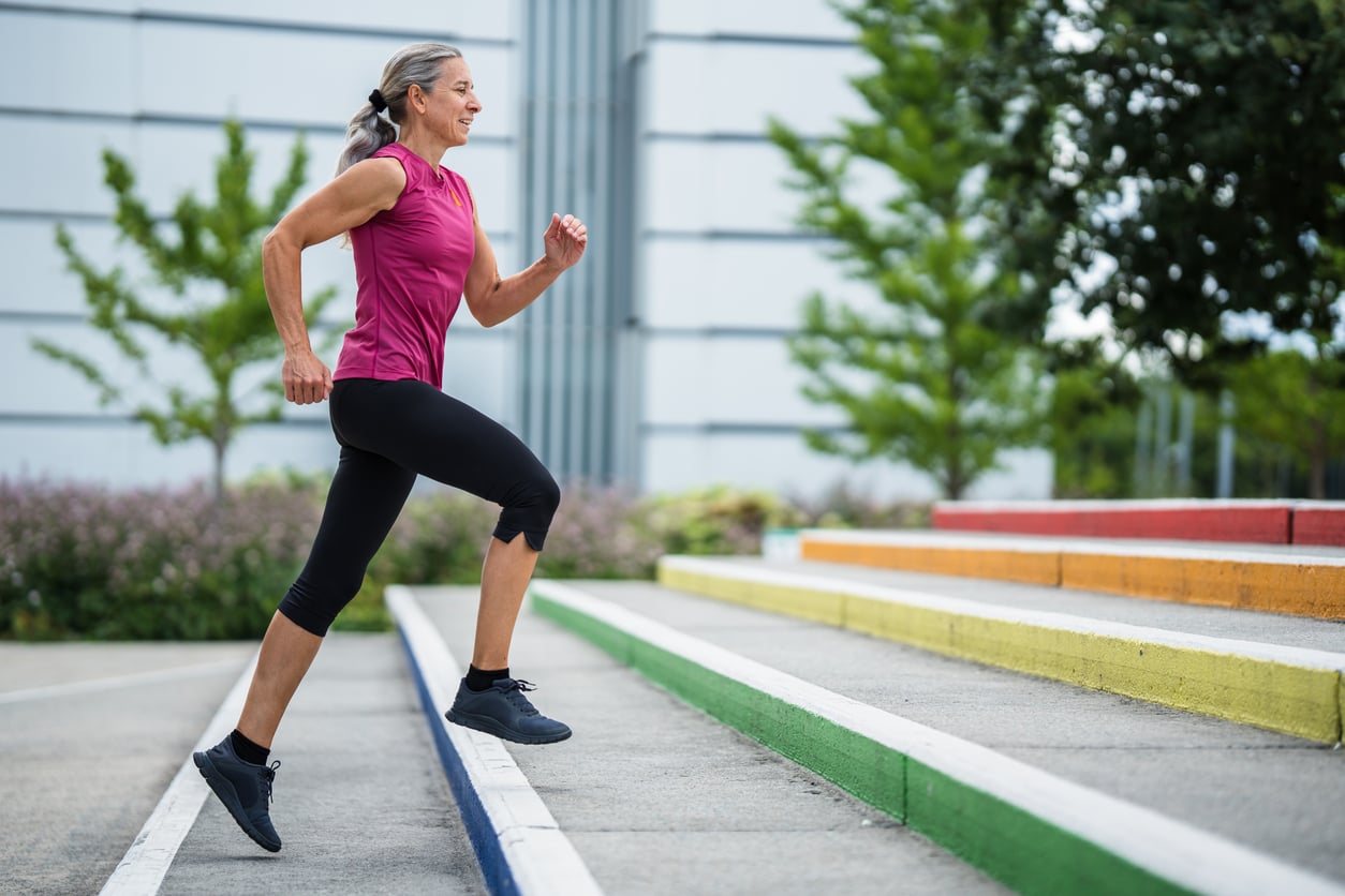 A woman in athletic clothing runs up colorful outdoor steps, exercising near a modern building with trees and plants in the background—a great way to stay fit and save money on gym fees.