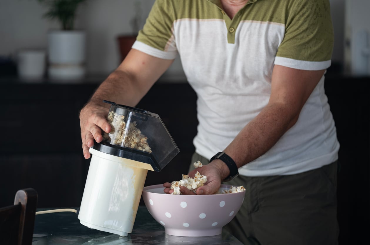 A person uses kitchen gadgets as they transfer fresh popcorn from an air popper into a pink bowl with white polka dots on a kitchen counter.