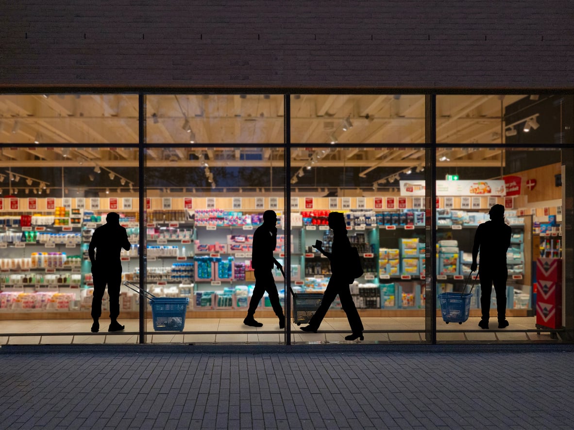 Silhouettes of four people with shopping baskets are seen through the window of a brightly lit supermarket, with shelves stocked with various products in the background.