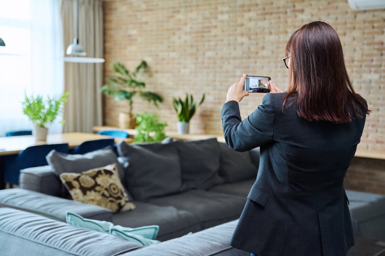 A person with long brown hair in a black blazer takes a photo of a modern living room with a gray sofa, green plants, and a brick wall, capturing stylish inspiration for anyone seeking hotel booking tips for their next trip.