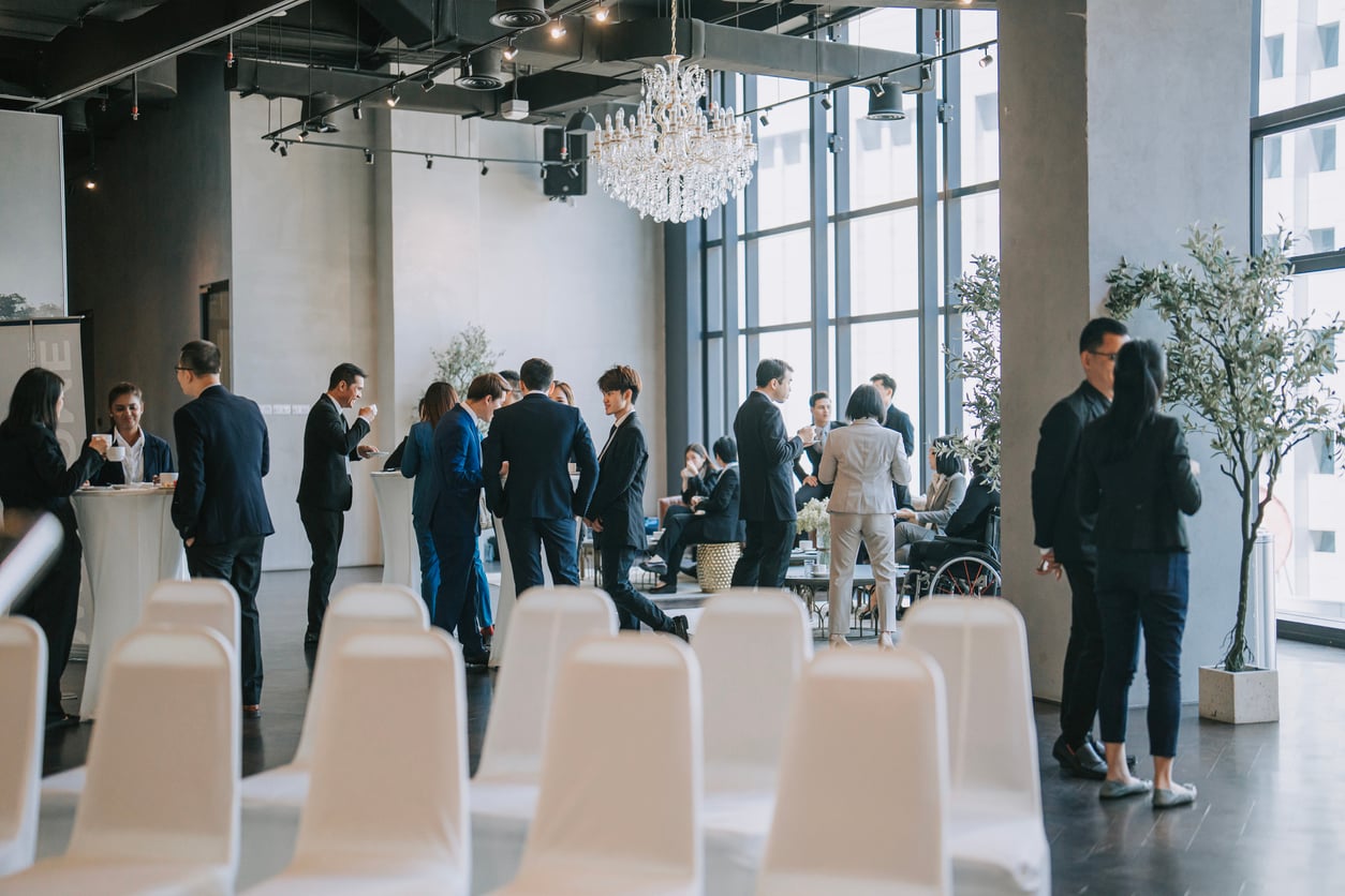 People in business attire mingle and converse in a modern, bright event space with large windows, a chandelier, white chairs, and plants—an ideal setting that leaves no room for common hotel booking mistakes.